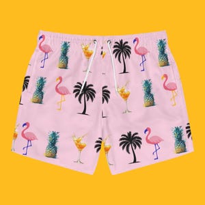 May include: Light pink swim shorts featuring an all-over print of pink flamingos, green pineapples, black palm tree silhouettes, and orange cocktails in stemmed glasses. The shorts have a white drawstring at the elasticated waistband.