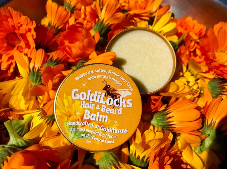 May include: A yellow tin of Goldilocks Hair & Beard Balm with a bee illustration. The tin is surrounded by orange and yellow calendula flowers.