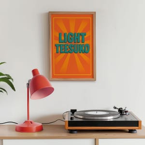 May include: A red desk lamp with a pink interior sits on a wooden surface next to a record player. A framed poster with the words "LIGHT TEESUKO" in teal letters against an orange background hangs on the wall.