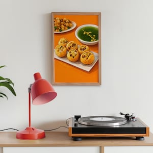 May include: A framed print depicting Indian street food: crispy, golden-brown puffed puri shells filled with savoury ingredients, served with green chutney and spiced potatoes. The print hangs above a red desk lamp and a record player.