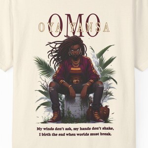 May include: Cream-colored t-shirt featuring a graphic of a person with dreadlocks, wearing a red jacket and black pants. The word "OMO" is printed above the graphic. Below the graphic is the text: "My winds don't ask, my hands don't shake, I birth the end when worlds must break."