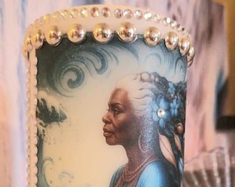 May include: A white candle featuring a colourful image of a woman with grey hair and a blue dress. The woman is seated in front of a blue wave. The candle is decorated with silver and white beads.
