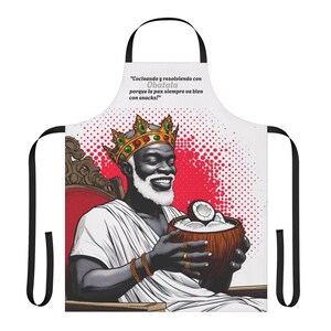 Peut inclure: Tablier blanc avec une illustration souriante d'un homme noir portant une couronne dorée et tenant une noix de coco. Le tablier a des bretelles noires et le texte "Cocinando y resolviendo con Obatala porque la paz siempre va bien con snacks!"