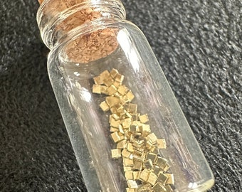 以下が含まれることがあります： コルク栓の付いた小さなガラス瓶に、小さな金色の金属の正方形が詰められています。
