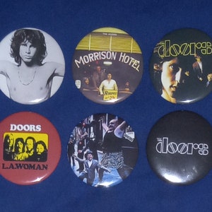 Könnte beinhalten: Sechs runde Anstecknadeln mit Motiven der Band The Doors. Die Nadeln zeigen verschiedene Bilder, darunter ein Schwarzweißfoto von Jim Morrison, ein Foto der Band vor einem Hotel und ein Foto von Jim Morrisons Gesicht. Die Nadeln zeigen auch den Bandnamen und den Albumtitel "LA Woman".