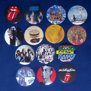 Könnte beinhalten: Eine Sammlung von Buttons der Band The Rolling Stones. Die Buttons zeigen Albumcover, das Bandlogo und andere Grafiken. Die Buttons sind rund und haben eine glänzende Oberfläche. Der Hintergrund ist einfarbig blau.