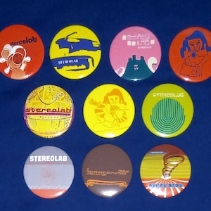 Puede incluir: Nueve botones redondos y coloridos con el nombre de la banda "Stereolab" impreso en ellos. Los botones presentan varios diseños, incluyendo un paisaje urbano, una mujer con un micrófono y un patrón geométrico.