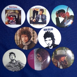 May include: A collection of eight round button badges featuring different images of Bob Dylan. The badges are white with black text and feature images of Bob Dylan from different eras of his career. The text on the badges includes "Bob Dylan", "Bringing It All Back Home", "Highway 61 Revisited", and "Bob Dylan".