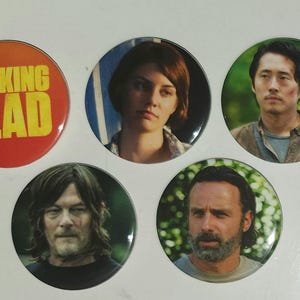 May include: A set of five circular pin-back buttons featuring The Walking Dead imagery. One button has a red background with yellow text reading The Walking Dead. The other four buttons display photographic portraits of four different characters.