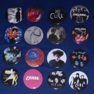 May include: A collection of 15 round The Cure band buttons. The buttons feature various album covers, band photos, and logos. The buttons are a mix of black, white, red, blue, and green.
