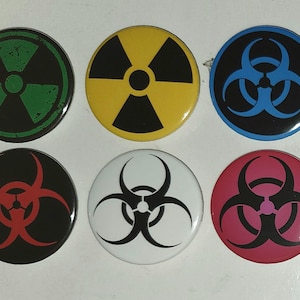 May include: A set of six circular pin-back buttons arranged in two rows. The top row features a green radiation symbol, a yellow radiation symbol, and a blue biohazard symbol. The bottom row displays a red biohazard symbol, a white biohazard symbol, and a pink biohazard symbol.