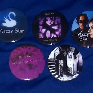 Op de afbeelding: Vijf ronde, kleurrijke knoppen met de bandnaam "Mazzy Star" erop gedrukt. De knoppen tonen verschillende albumhoezen en bandfoto's.