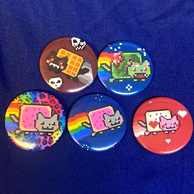 May include: Six round, colorful, pinback buttons featuring a pixelated cat character with different backgrounds. The cat is wearing a pink slice of bread with a smiling face. The backgrounds include a rainbow, a leopard print, a red background with hearts, a blue background with stars, a brown background with skulls, and a green background with a rainbow.