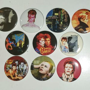 Może przedstawiać: Kolekcja dziesięciu okrągłych przypinek z różnymi kultowymi okładkami albumów i portretami Davida Bowiego. Wzory obejmują grafiki z Ziggy Stardust, Low, Diamond Dogs i Hunky Dory, prezentując różnorodne palety kolorów i style artystyczne.
