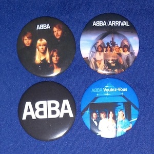 Peut inclure: Quatre boutons ronds noirs et blancs avec des images du groupe ABBA. Les boutons présentent le nom du groupe et les pochettes d'album pour "Arrival", "Voulez-Vous" et "ABBA".
