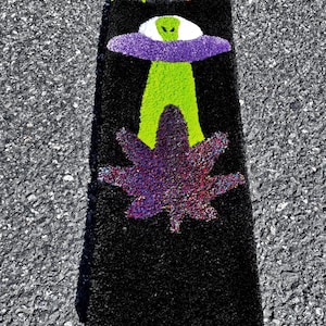 May include: Black rectangular rug with a lighter-shaped design. The rug features a green alien with a purple UFO, and a purple and red cannabis leaf. The top of the rug has a red and green design.