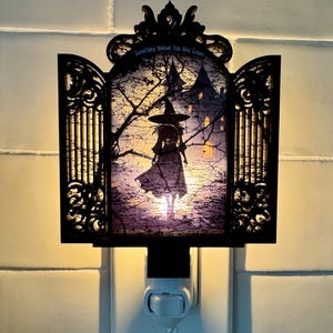 May include: Black night light with a witch silhouette in front of a castle. The night light has a decorative black frame with the text "Something Wicked This Way Comes". The night light plugs into a standard wall outlet.