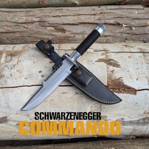 May include: A silver knife with a black handle and a leather sheath. The knife has a serrated edge and a pointed tip. The sheath has a brown leather strap with a metal buckle. The knife is lying on a wooden surface. The text "SCHWARZENEGGER COMMANDO" is printed on the wooden surface.