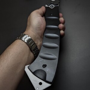 May include: A matte black tactical axe with a curved handle featuring four distinct oval indentations for grip. The axe head is made of brushed silver metal with a single circular hole near the bottom edge and a visible bolt near the handle base.