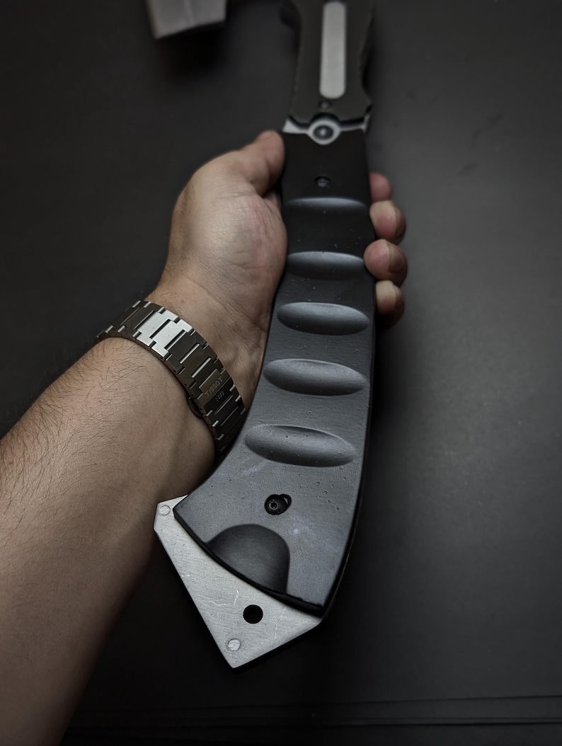 May include: A matte black tactical axe with a curved handle featuring four distinct oval indentations for grip. The axe head is made of brushed silver metal with a single circular hole near the bottom edge and a visible bolt near the handle base.