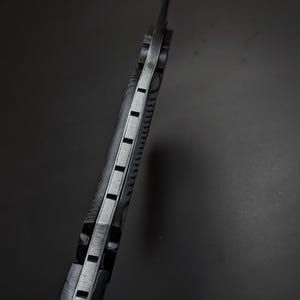 May include: A side view of a knife handle featuring a metallic tang sandwiched between two textured black grip scales. The spine of the handle displays a series of evenly spaced rectangular notches cut into the metal, creating a distinct pattern.