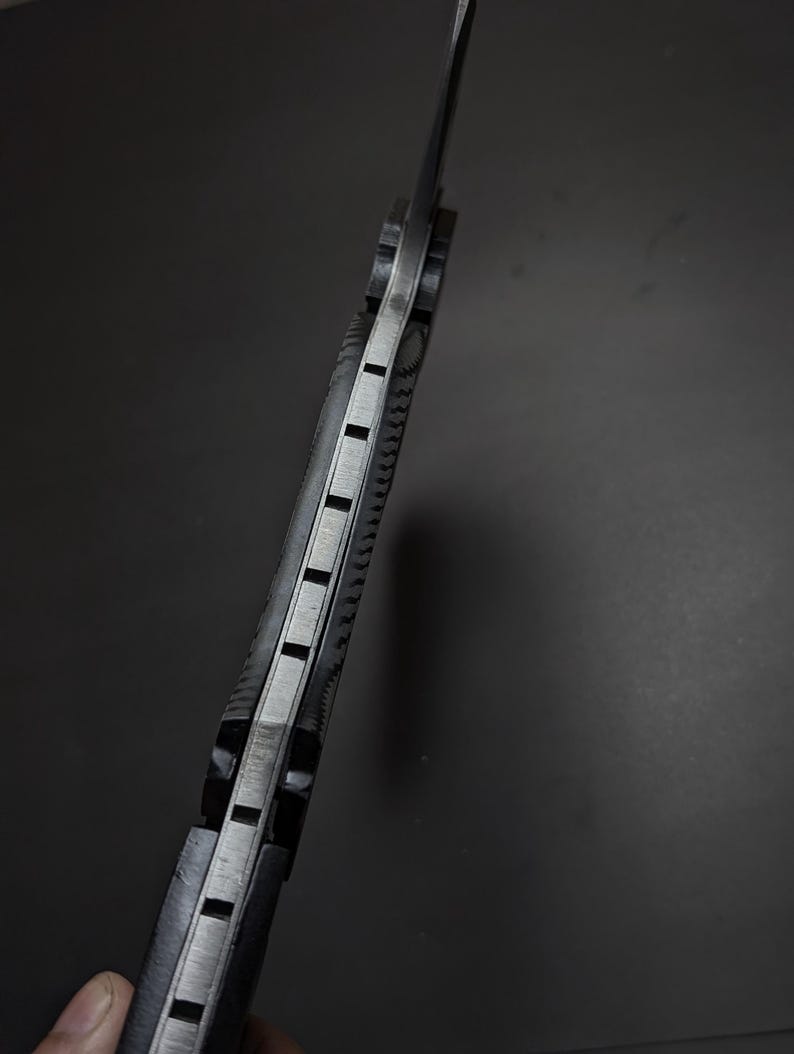 May include: A side view of a knife handle featuring a metallic tang sandwiched between two textured black grip scales. The spine of the handle displays a series of evenly spaced rectangular notches cut into the metal, creating a distinct pattern.