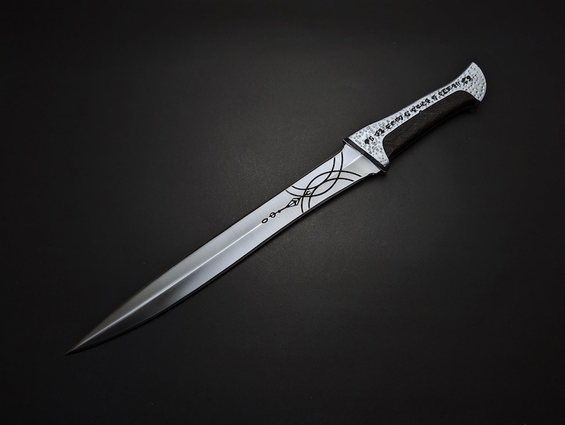 K&ouml;nnte beinhalten: Ein silberner Dolch mit spitzer Klinge und dekorativem Griff. Der Griff hat einen schwarzen Griff und wei&szlig;e Akzente mit ge&auml;tzten Mustern und Text. Die Klinge hat ge&auml;tzte Designs in der N&auml;he des Griffs.