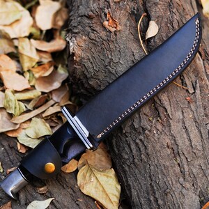 May include: A black leather knife sheath with a brown stitching detail. The sheath is lying on a tree trunk with fallen leaves.