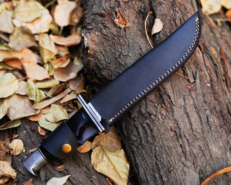 May include: A black leather knife sheath with a brown stitching detail. The sheath is lying on a tree trunk with fallen leaves.