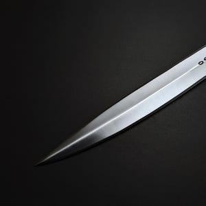 K&ouml;nnte beinhalten: Nahaufnahme eines silbernen Dolches mit scharfer Spitze. Die Klinge hat ein dekoratives ge&auml;tztes Design in der N&auml;he des Griffs. Der Dolch ist vor einem dunklen Hintergrund platziert, der seinen metallischen Glanz hervorhebt.
