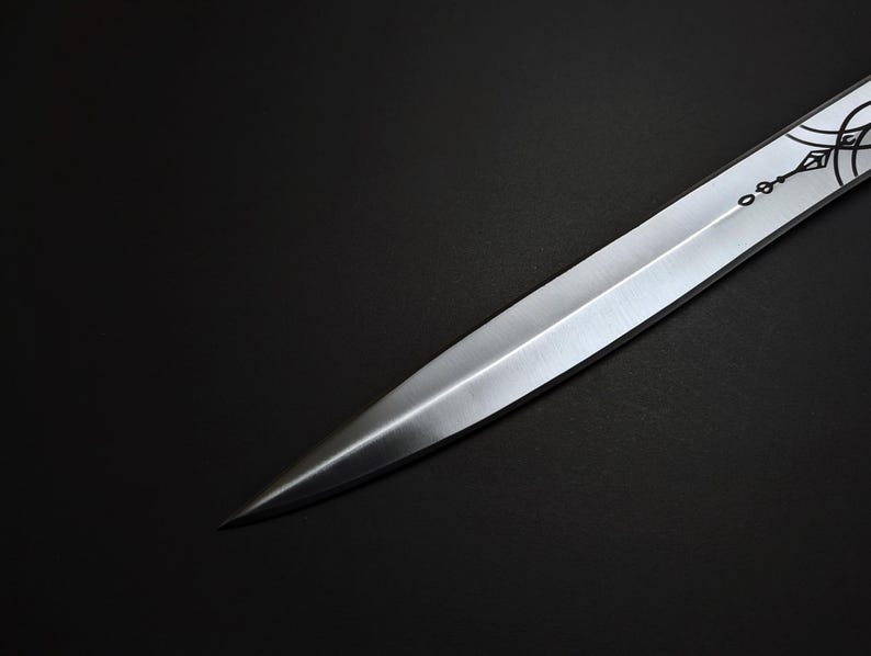 K&ouml;nnte beinhalten: Nahaufnahme eines silbernen Dolches mit scharfer Spitze. Die Klinge hat ein dekoratives ge&auml;tztes Design in der N&auml;he des Griffs. Der Dolch ist vor einem dunklen Hintergrund platziert, der seinen metallischen Glanz hervorhebt.