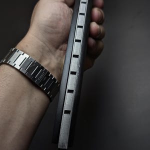 May include: A silver-toned Tissot 1853 watch with a metallic link bracelet and a black rectangular object featuring a series of evenly spaced, recessed silver-colored square indentations along its side edge, held in a vertical orientation.