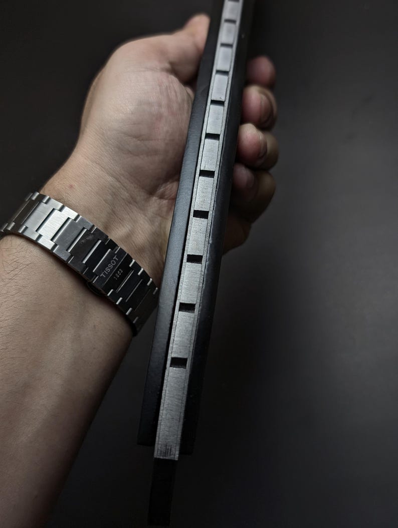 May include: A silver-toned Tissot 1853 watch with a metallic link bracelet and a black rectangular object featuring a series of evenly spaced, recessed silver-colored square indentations along its side edge, held in a vertical orientation.