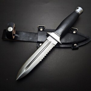 May include: A stainless steel dagger with a serrated edge and black handle rests on a black leather sheath. The dagger has a silver bolster and a pointed tip. The sheath has a belt loop.