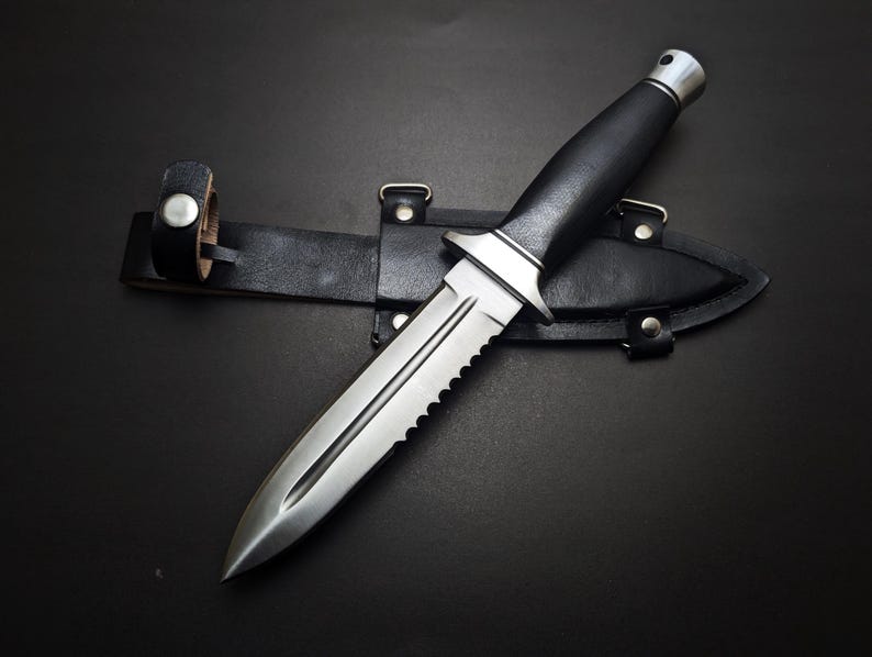May include: A stainless steel dagger with a serrated edge and black handle rests on a black leather sheath. The dagger has a silver bolster and a pointed tip. The sheath has a belt loop.