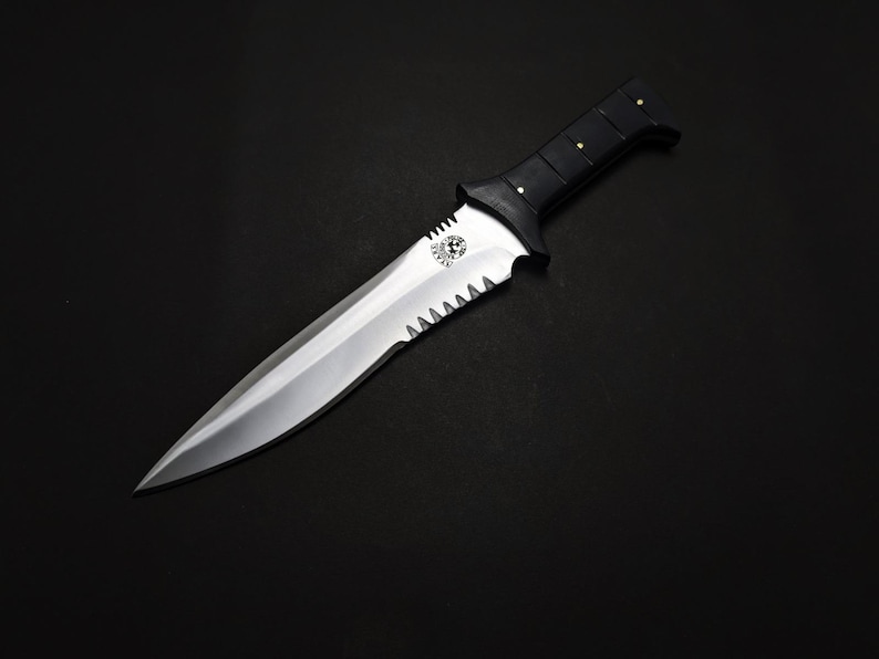 May include: A silver and black knife with a serrated edge and a pointed tip. The handle is black with gold-colored rivets. The knife has a decorative emblem near the blade's base. The background is black.