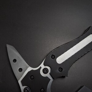 May include: A tactical tool featuring a sharp, pointed metal blade with two circular cutouts. The handle is constructed with black textured grip panels secured by multiple small screws, featuring a central silver metallic inlay along the spine.
