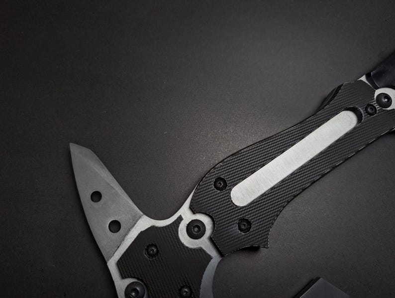 May include: A tactical tool featuring a sharp, pointed metal blade with two circular cutouts. The handle is constructed with black textured grip panels secured by multiple small screws, featuring a central silver metallic inlay along the spine.