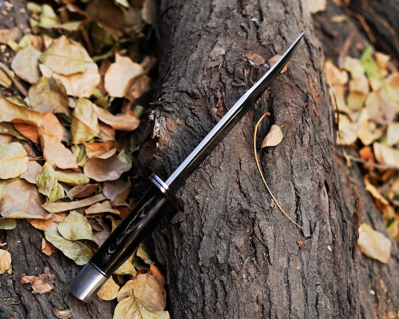 May include: A black-handled hunting knife with a silver blade, lying on a tree trunk covered in brown bark and surrounded by fallen leaves.