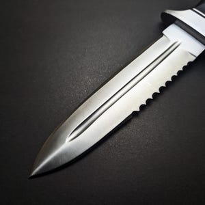 May include: A close-up of a stainless steel dagger with a serrated edge and a black handle. The blade has a central groove and a sharp point. The dagger is set against a dark background.