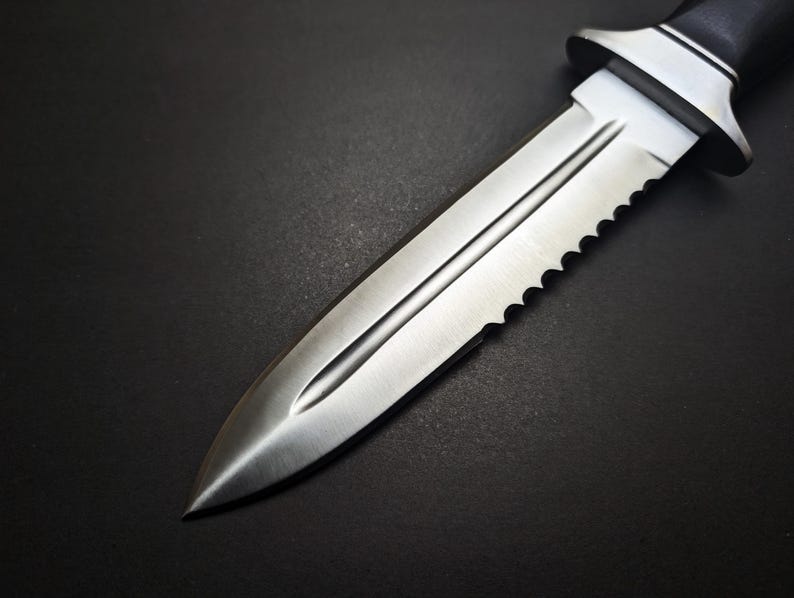 May include: A close-up of a stainless steel dagger with a serrated edge and a black handle. The blade has a central groove and a sharp point. The dagger is set against a dark background.
