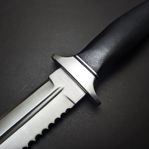 May include: Close-up of a silver and black knife. The blade is partially serrated and has a sharp point. The handle is black with a textured surface. The knife has a silver guard and a silver end cap.