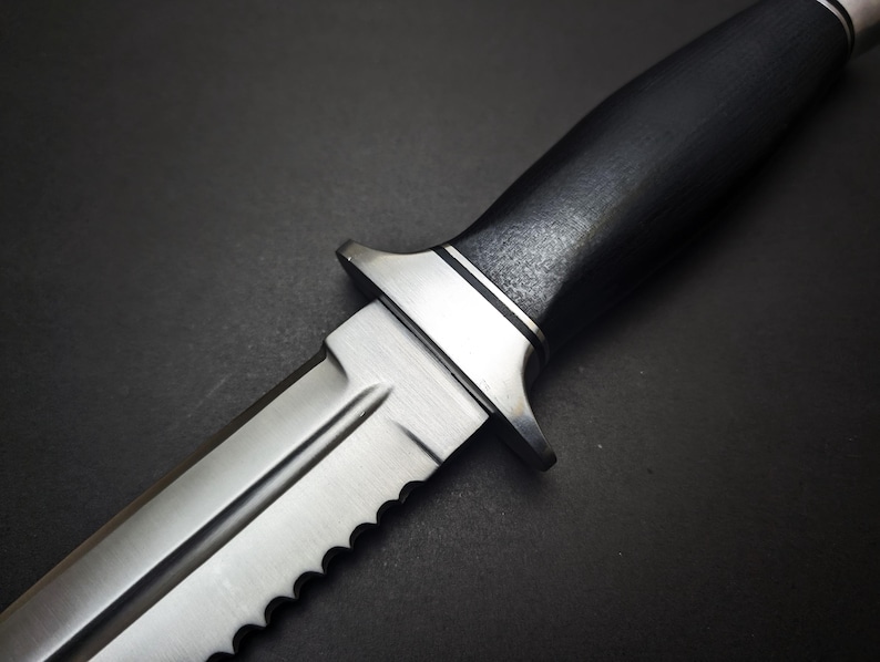 May include: Close-up of a silver and black knife. The blade is partially serrated and has a sharp point. The handle is black with a textured surface. The knife has a silver guard and a silver end cap.