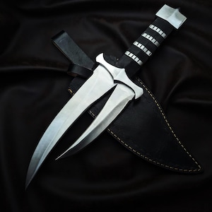 May include: A silver and black dagger with a unique forked blade design. The handle is black with silver accents, and it rests in a black leather sheath. The dagger is displayed against a black background.