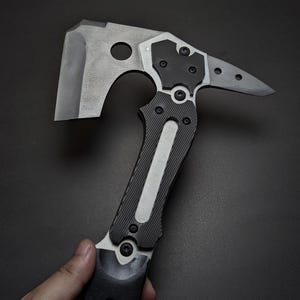 May include: A tactical hatchet featuring a matte grey metal axe head with a circular cutout, a pointed rear spike, and a black textured handle secured with multiple visible hex screws. The tool has a modern industrial design with metallic accents.