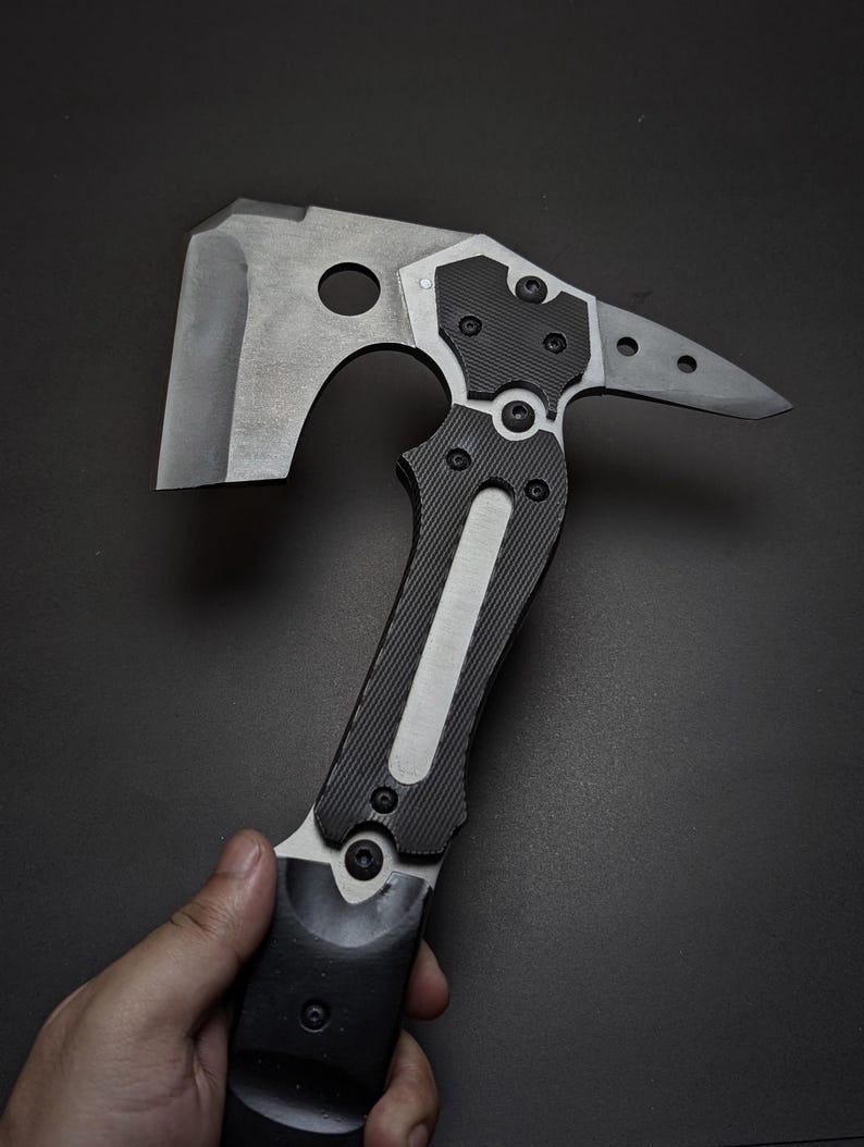 May include: A tactical hatchet featuring a matte grey metal axe head with a circular cutout, a pointed rear spike, and a black textured handle secured with multiple visible hex screws. The tool has a modern industrial design with metallic accents.