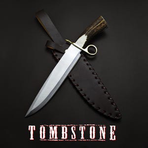 May include: A large, silver-bladed knife with a brown antler handle and a leather sheath. The knife is resting on a black surface, with the word "TOMBSTONE" in red and white text at the bottom.