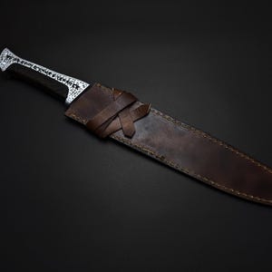 K&ouml;nnte beinhalten: Ein handgefertigtes Messer mit dunklem Griff und dekorativer silberner Klinge, in einer braunen Lederscheide. Die Scheide hat einen Verschluss aus geflochtenem Lederriemen. Messer und Scheide sind vor dunklem Hintergrund platziert.