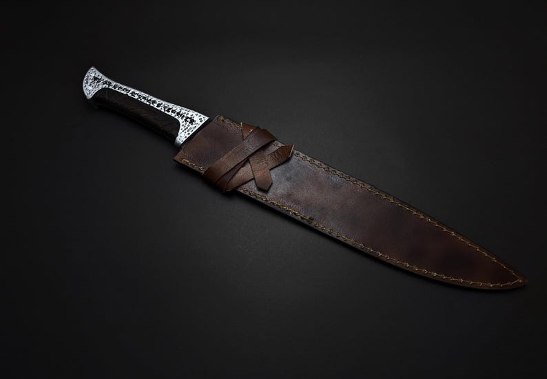 K&ouml;nnte beinhalten: Ein handgefertigtes Messer mit dunklem Griff und dekorativer silberner Klinge, in einer braunen Lederscheide. Die Scheide hat einen Verschluss aus geflochtenem Lederriemen. Messer und Scheide sind vor dunklem Hintergrund platziert.