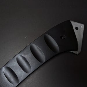 May include: A black handle with a curved shape and four distinct oval indentations along its length. The handle features a metallic silver-toned end piece secured with a small black screw, showcasing a sleek and functional design for a tool.